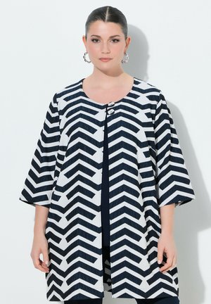 Kvinde med mørkt hår i en lav knold bærer en sort og hvid chevron-mønstret lang jakke med trekvartærmer og sølvfarvede øreringe i hoop-stil.