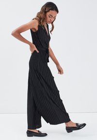 Jumpsuit noir plissé avec un décolleté en V profond, ceinture à la taille et jambes larges. Porté avec des mocassins noirs présentant un accent décoratif.