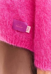 Jasnoróżowy, puszysty materiał swetra o tkanej fakturze. Zawiera fioletową metkę z napisem „Knitwear” oraz mały różowy akcent.