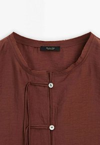 Chemise en lin marron avec encolure ronde, deux boutons blancs et détails à nouer sur la patte de boutonnage avant, étiquette indiquant "Massimo Dutti Fabriqué au Portugal."