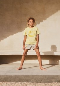 Gul t-skjorte med frukt-smiley trykk, kombinert med grå shorts. Begge plaggene har avslappet passform og myk tekstur. Barnet står barføtt.