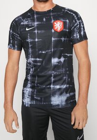 Camiseta deportiva negra de manga corta con un patrón texturizado de tie-dye en gris. Presenta un emblema de león rojo y el logo de Nike en el pecho.