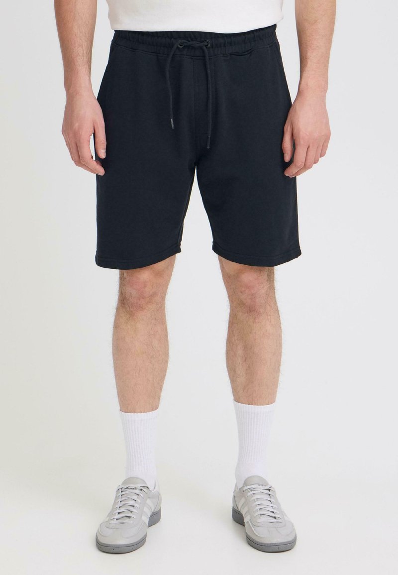 Homme debout portant un short noir à cordon, des chaussettes blanches montantes, des baskets grises et une chemise blanche sur un fond uni.