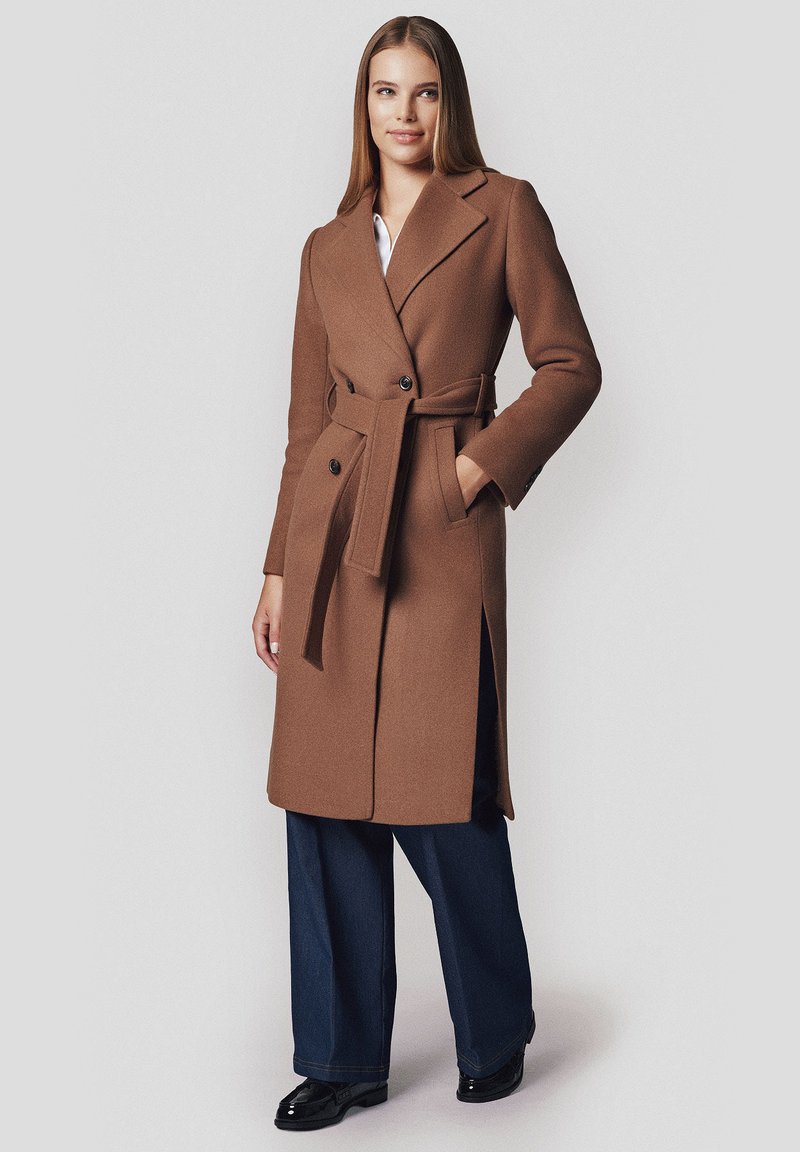 Brązowy wełniany płaszcz o dopasowanym kroju, z szerokimi klapami, podwójnym zapięciem na guziki i pasującym paskiem; zestawiony z szerokimi spodniami dżinsowymi i butami.