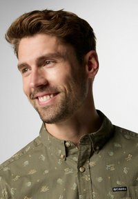 Camicetta verde con colletto, caratterizzata da motivi botanici blu chiaro e gialli. Include una piccola etichetta con il logo Columbia.