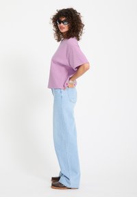 Frau mit lockigem Haar, die schwarze Sonnenbrille, ein lila T-Shirt, hellblaue weite Jeans, Armbänder und braune Mokassins trägt und seitlich steht.