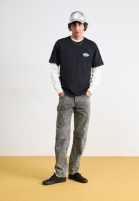 Camiseta de algodón negra superpuesta sobre una camisa de manga larga blanca, combinada con unos jeans de mezclilla gris y zapatillas negras. Una gorra plateada completa el conjunto.