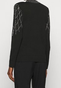 Czarny top z długim rękawem z wzorami w kształcie rombów z kryształkami na rękawach i kołnierzu, noszony z pasującymi czarnymi spodniami z tyłu.