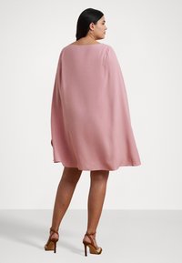 Vestido rosa con capa, escote cuadrado, mangas cortas abullonadas y una silueta fluida. Combinado con sandalias de tacón alto. Textura de tela suave.