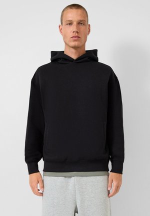 Mann mit kurzem Haar trägt einen schwarzen Hoodie und hellgraue Jogginghosen, steht vor einem einfarbigen hellgrauen Hintergrund.