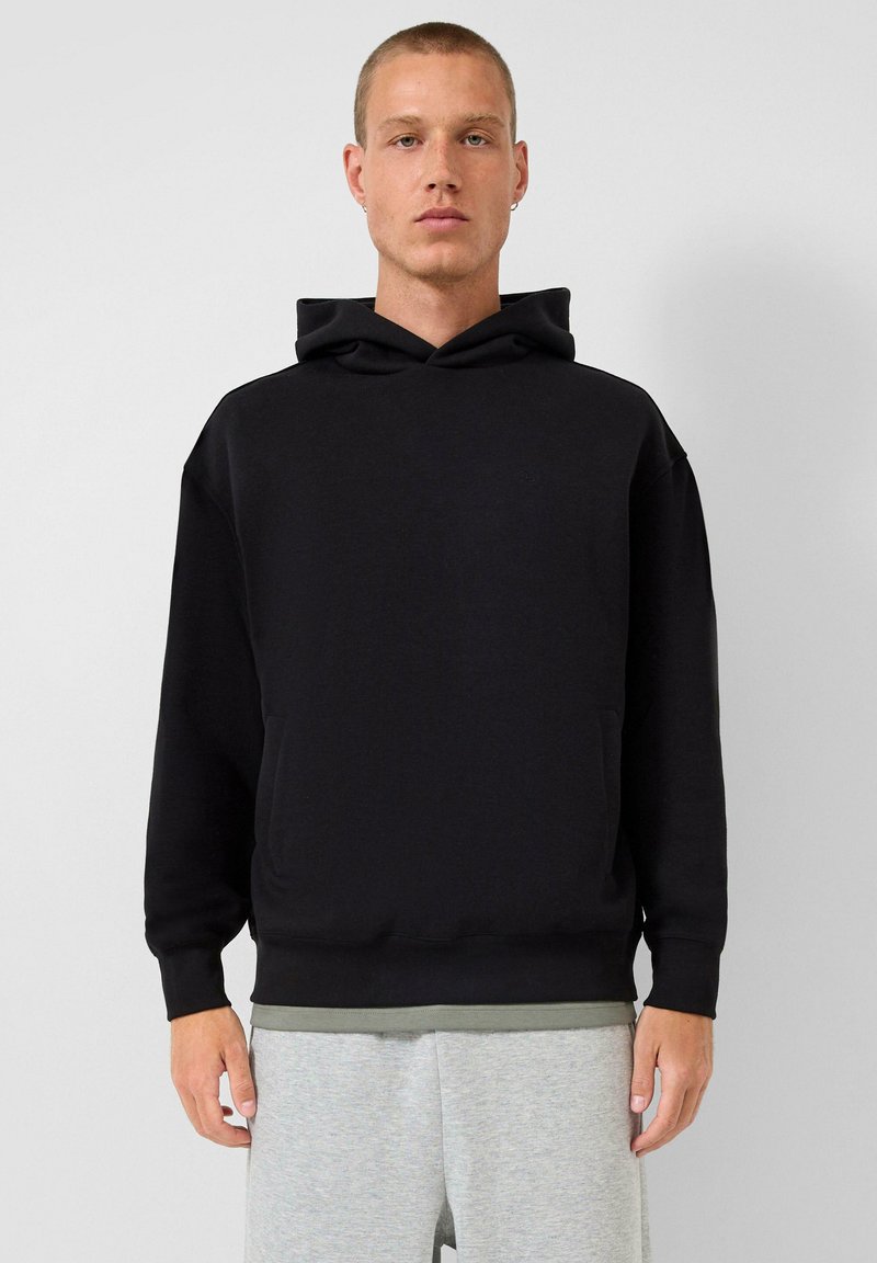 Uomo con capelli corti, indossa un hoodie nero e pantaloni grigi chiari, in piedi contro uno sfondo grigio chiaro uniforme.