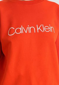 Červená mikina z měkké textilie s bílým logem "Calvin Klein" přes hrudník. Kulatý výstřih a volný střih.