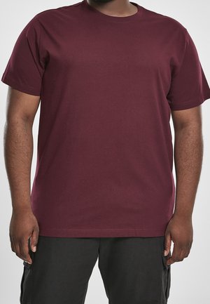 Hombre con camiseta de manga corta lisa color granate y pantalones negros, mostrado desde el cuello hasta la mitad del muslo sobre un fondo blanco.