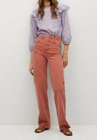 Helles lila Bluse mit Blumenmuster und Rüschensleeves, kombiniert mit hoch taillierten, staubigen rosa weiten Hosen. Braune Sandalen ergänzen das Outfit.