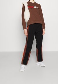 Sudadera de forro polar marrón con un logotipo rojo y blanco, combinada con pantalones negros que presentan rayas laterales marrones y naranjas. Zapatillas blancas completan el conjunto.
