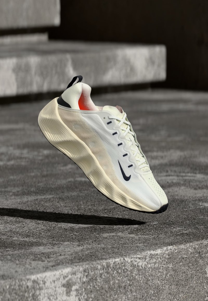 Chaussure de course Nike blanche et beige avec semelle épaisse et logo Swoosh noir, flottant au-dessus d'une surface en béton avec des marches en arrière-plan.