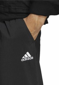 Schwarze Sporthosen mit elastischem Bund, versehen mit einem weißen gestickten Adidas-Logo auf der unteren linken Seite. Das Material wirkt leicht.