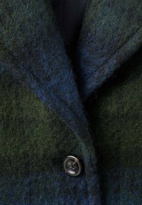 Cappotto in misto lana con superficie testurizzata in tonalità di verde scuro e blu, con un evidente dettaglio dei bottoni e un design con lapels a punta.