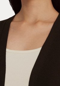 Cardigan a coste marrone scuro indossato sopra un top a coste beige chiaro, che mostra materiali morbidi e testurizzati e un design con scollo a V.