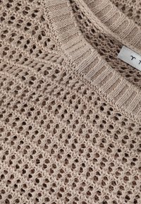 Maglione lavorato a maglia in beige, con un motivo a trama aperta, scollatura a coste e tessuto testurizzato. Etichetta visibile vicino al colletto.