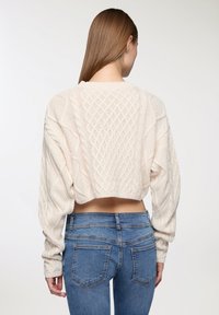 Kremowy krótki sweter z wzorem w warkocze i objętościowymi rękawami, w parze z niebieskimi obcisłymi dżinsami, widok z tyłu.