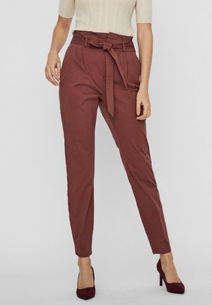 Rostfarbene, hoch taillierte Hose mit Stoffgürtel, zulaufendem Bein und glatter Textur. Kombiniert mit burgunderroten Absätzen.