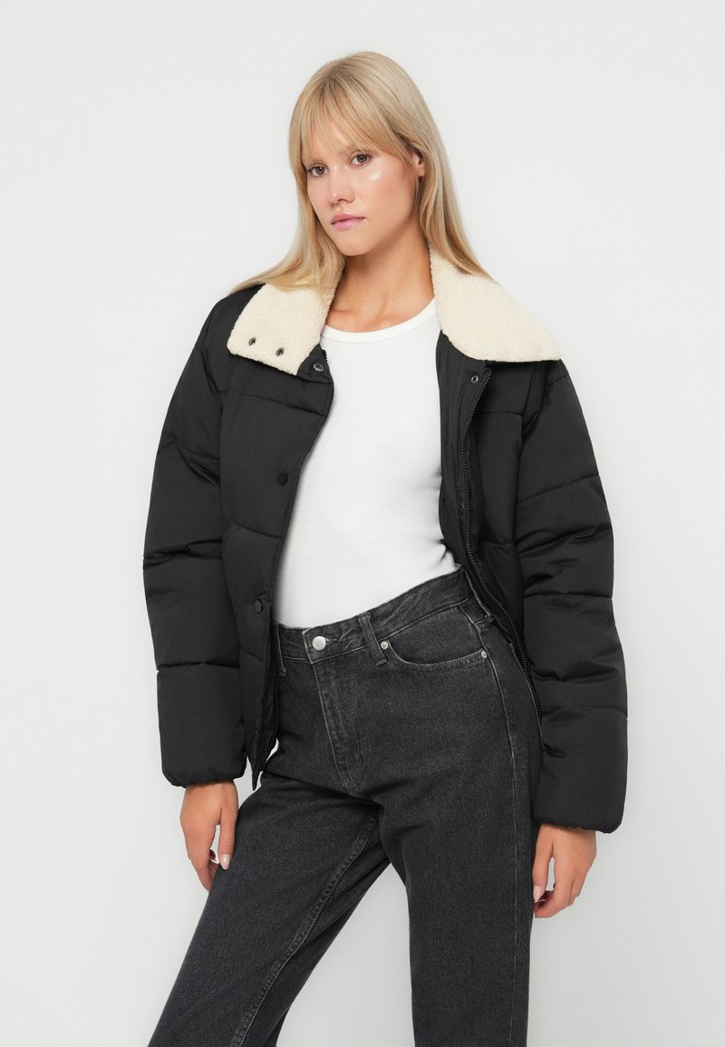 Veste matelassée noire avec un col en fausse toison crème, dotée d'une fermeture éclair et de boutons-pression. Portée par-dessus une chemise blanche et un jean foncé.