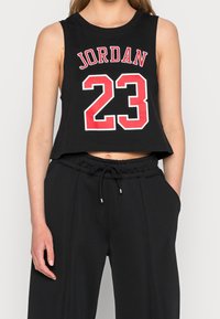 Musta lyhyt toppi pyöreällä kaula-aukolla, jossa punainen ja valkoinen "JORDAN"-teksti ja numero "23". Yhdistetty löysiin mustiin collegehousuihin.