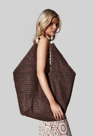 Donna con capelli biondi che porta un grande borsone intrecciato marrone scuro con manici decorati con perline, indossando un vestito bianco all'uncinetto, su uno sfondo grigio semplice.