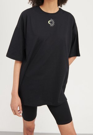 Persoon draagt een oversized zwart t-shirt met een kleine afbeelding van een halvemaan en zon in het midden van de borst, gecombineerd met zwarte fietsshorts.