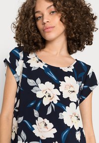 Top floral azul marinho feito de tecido leve. Apresenta um decote redondo, mangas curtas e grandes flores cor-de-rosa com folhas azul-esverdeadas.