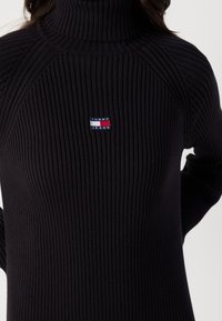 Pull à col roulé noir en tricot côtelé avec un petit patch rectangulaire du logo Tommy Jeans centré sur la poitrine.