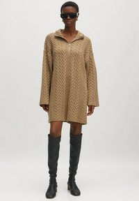 Beiges Strickkleid mit einem Frontreißverschluss und großem Kragen. Lange Ärmel mit gerippten Bündchen, kombiniert mit knielangen schwarzen Stiefeln.