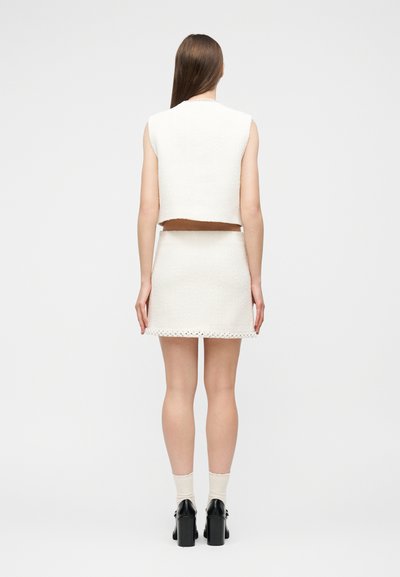 Femme aux longs cheveux bruns portant un haut sans manches blanc texturé et une jupe assortie, chaussures à talons noires, debout devant un fond uni.