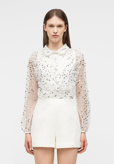Femme portant un short blanc taille haute et un chemisier transparent à manches longues orné de sequins noirs et argentés ainsi que de boutons en perle.