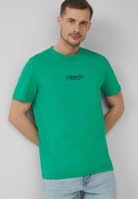 Groene katoenen T-shirt met korte mouwen, ronde halslijn en zwart "s.Oliver Co. Established 1969" logo op de voorkant. Zachte textuur.