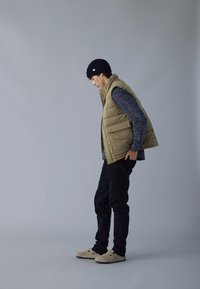 Joven que lleva un gorro negro, un chaleco acolchado beige, un sudadera gris, pantalones negros y zapatos beiges sin cordones, de pie y mirando hacia abajo.
