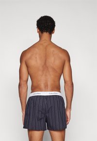 Melni svītraini bokseri ar baltu Calvin Klein jostu, piedāvājot brīvu piegriezumu un gludu auduma tekstūru. Produktu skatījums no aizmugures.