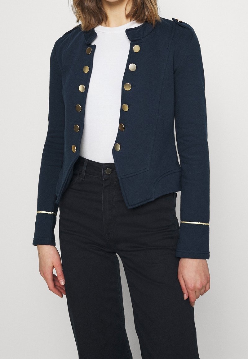Personne portant une veste bleu marine de style militaire avec des boutons dorés, une chemise blanche et un pantalon noir, recadrée pour ne montrer que le torse et les bras.