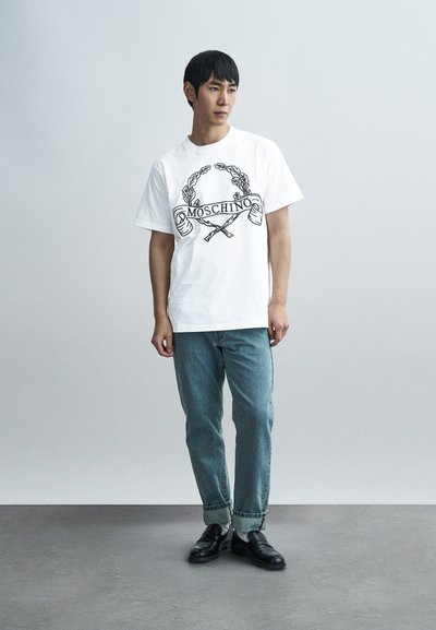 Λευκό βαμβακερό μπλουζάκι με μαύρο γραφικό τύπωμα που γράφει "Moschino" και διακοσμητικά στοιχεία, σε συνδυασμό με ανοιχτό μπλε τζιν παντελόνι και μαύρα παπούτσια.