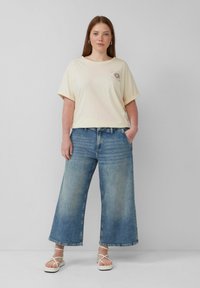 Beige übergroßes T-Shirt mit kurzen Ärmeln und Sonnenmotiv; hellblaue weite Jeans; cremefarbene Sandalen mit Riemchen; Model steht mit einer Hand in der Tasche.