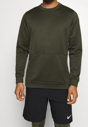 Mann trägt ein olivgrünes Sweatshirt mit langen Ärmeln und vorderer Reißverschlusstasche sowie schwarze Nike-Shorts mit weißem Swoosh-Logo.