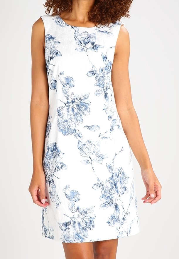 Vestido blanco sin mangas con estampado floral azul llevado por una persona con cabello rizado, de pie frente a un fondo blanco uniforme.