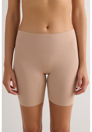 Gros plan d'une personne portant un cuissard beige taille haute sans coutures, montrant la section médiane et le haut des jambes sur un fond uni.