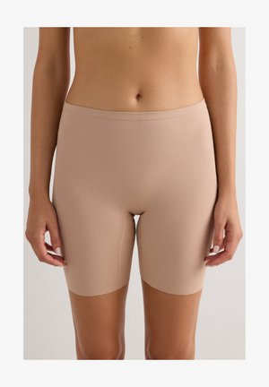 Nahaufnahme einer Person, die beige nahtlose Biker-Shorts mit hoher Taille trägt, und dabei den Mittelteil sowie die oberen Beine vor einem einfarbigen Hintergrund zeigt.