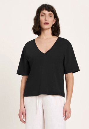 Frau mit kurzen dunklen Haaren, die ein schwarzes V-Ausschnitt-Kurzarmshirt und lockere weiße Hose trägt und vor einem einfarbigen Hintergrund steht.
