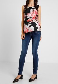 Mujer vestida con una blusa sin mangas negra con un gran estampado floral rosa, jeans ajustados azul oscuro y zapatos negros de tacón bajo, de pie contra un fondo liso.