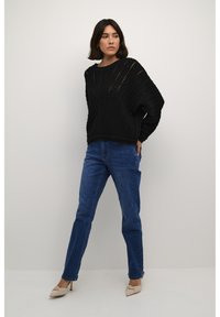 Svart stickad tröja med en texturerad, öppen mönster och nedhasade axlar, tillsammans med blå raka jeans och beige spetsiga skor.