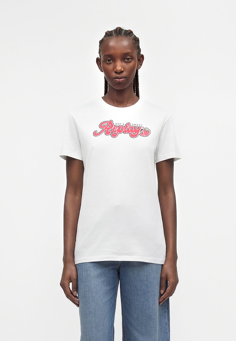 Γυναίκα με πλεξούδες που φοράει λευκό μπλουζάκι με ροζ κείμενο "Replay Hot & Sweet" και γραφικό φράουλας, σε συνδυασμό με μπλε τζιν.