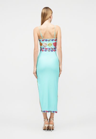 Kvinde med langt blondt hår iført en lyseblå, ankellang kjole med spaghetti-stropper og farverig blomsterbroderi på toppen og nederst, stående.